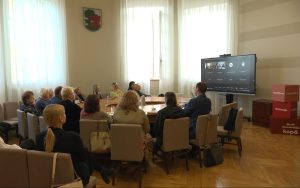 Cilvēki piedalās diskusijā pašvaldības zālē, daļa dalībnieku pieslēgušies tiešsaistē