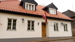 Gaiša ēka ar sarkanu dakstiņu jumtu, Latvijas karogu un uzrakstu “Spīdalas nams”.