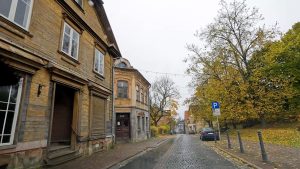 Klusa bruģēta iela ar vecpilsētas ēkām un kokiem rudens krāsās.