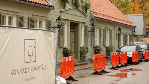 Atjaunota zaļgani pelēka koka ēka ar sarkanu dakstiņu jumtu un oranžiem drošības barjeriem uz ielas.