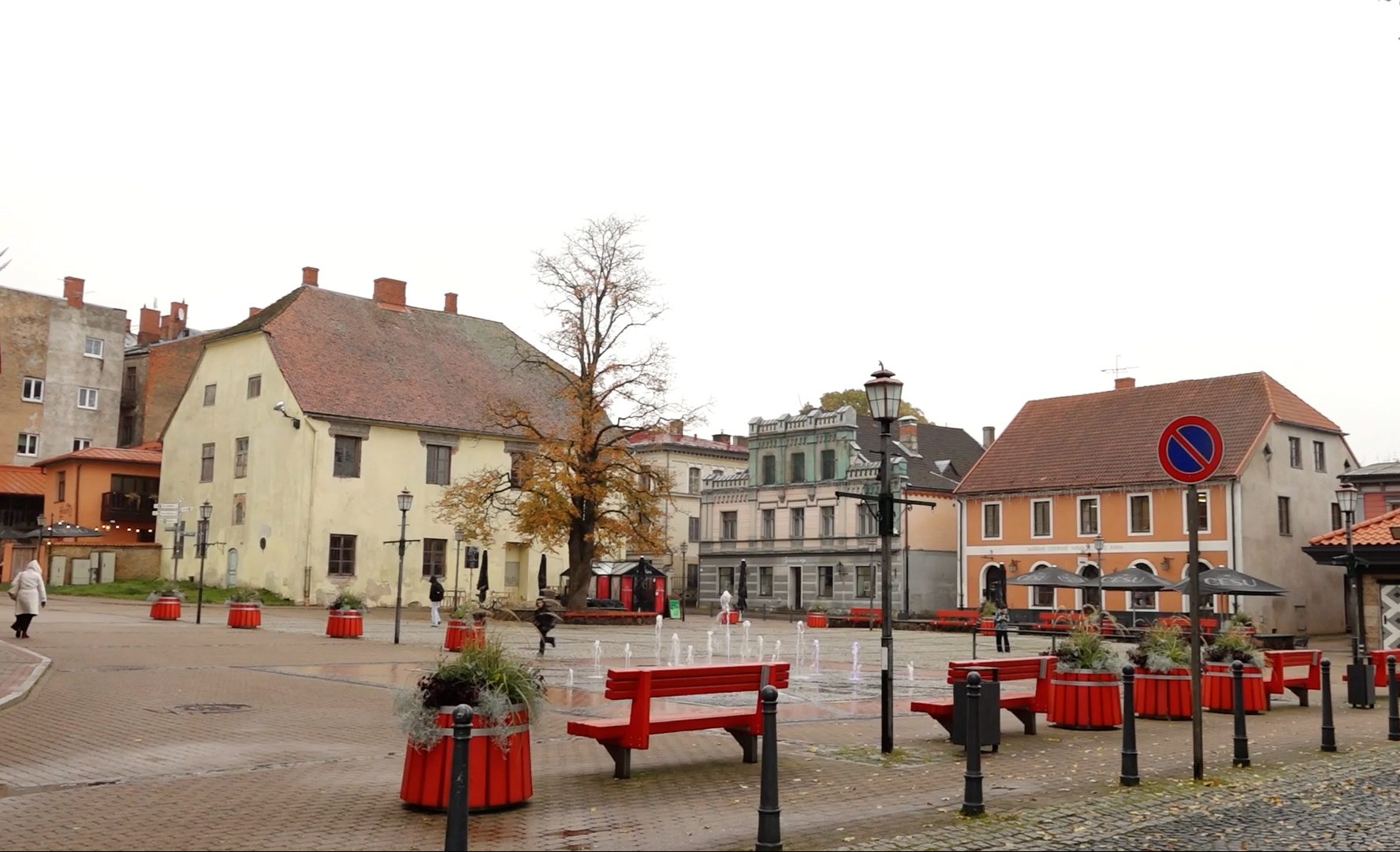 Klusa pilsētas laukuma ainava ar vēsturiskām ēkām un sarkanām soliņām.