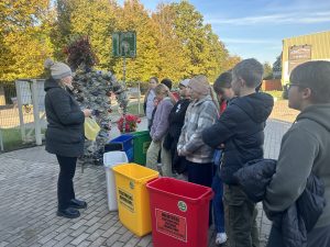 Skolēni piedalās vides izglītības nodarbībā par atkritumu šķirošanu ārā pie konteineriem.