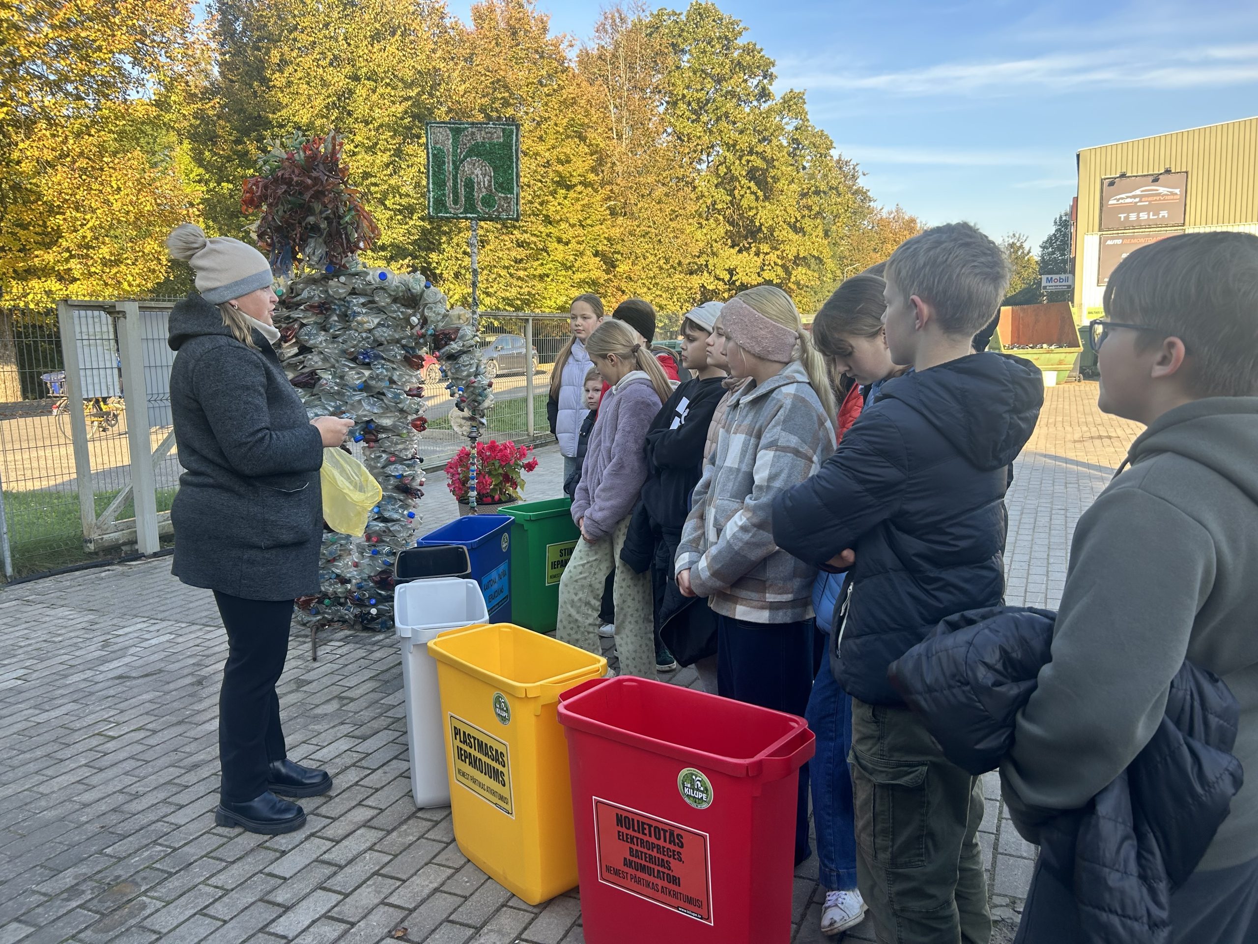 Skolēni piedalās vides izglītības nodarbībā par atkritumu šķirošanu ārā pie konteineriem.