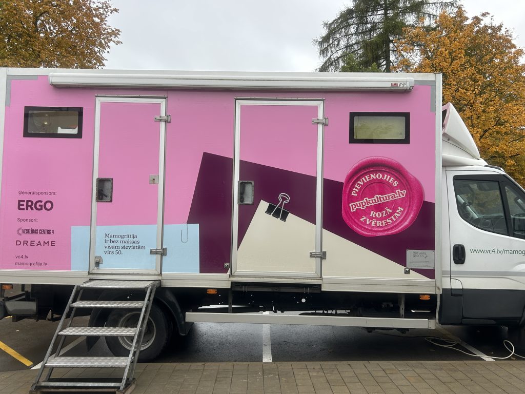Mobilais mamogrāfa furgons spilgti rozā krāsā, novietots pie ģimenes ārsta prakses Ikšķilē, ar metāla kāpnēm un informāciju par skrīningu.