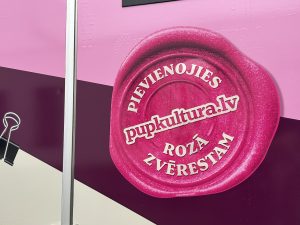 Tuvplānā redzams uzlīmēts rozā zīmoga attēls uz mamogrāfa furgona, ar uzrakstu "PIEVIENOJIES pupkultura.lv ROZĀ ZVĒRESTAM".