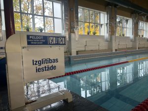 Skats uz peldbaseinu ar uzrakstu “Izglītības iestāde” un norādi “Peldot, lūdzu, turies celiņa labajā pusē”.