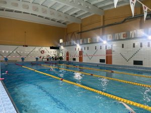 Peldbaseins ar peldētājiem treniņa laikā, redzamas peldceliņu līnijas un sporta simboli uz sienām.