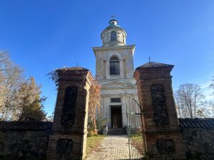 Skats uz Sunākstes (Stendera) baznīcas ieeju caur veciem ķieģeļu vārtiem ar metāla režģi, kas atrodas zem augsta torņa.