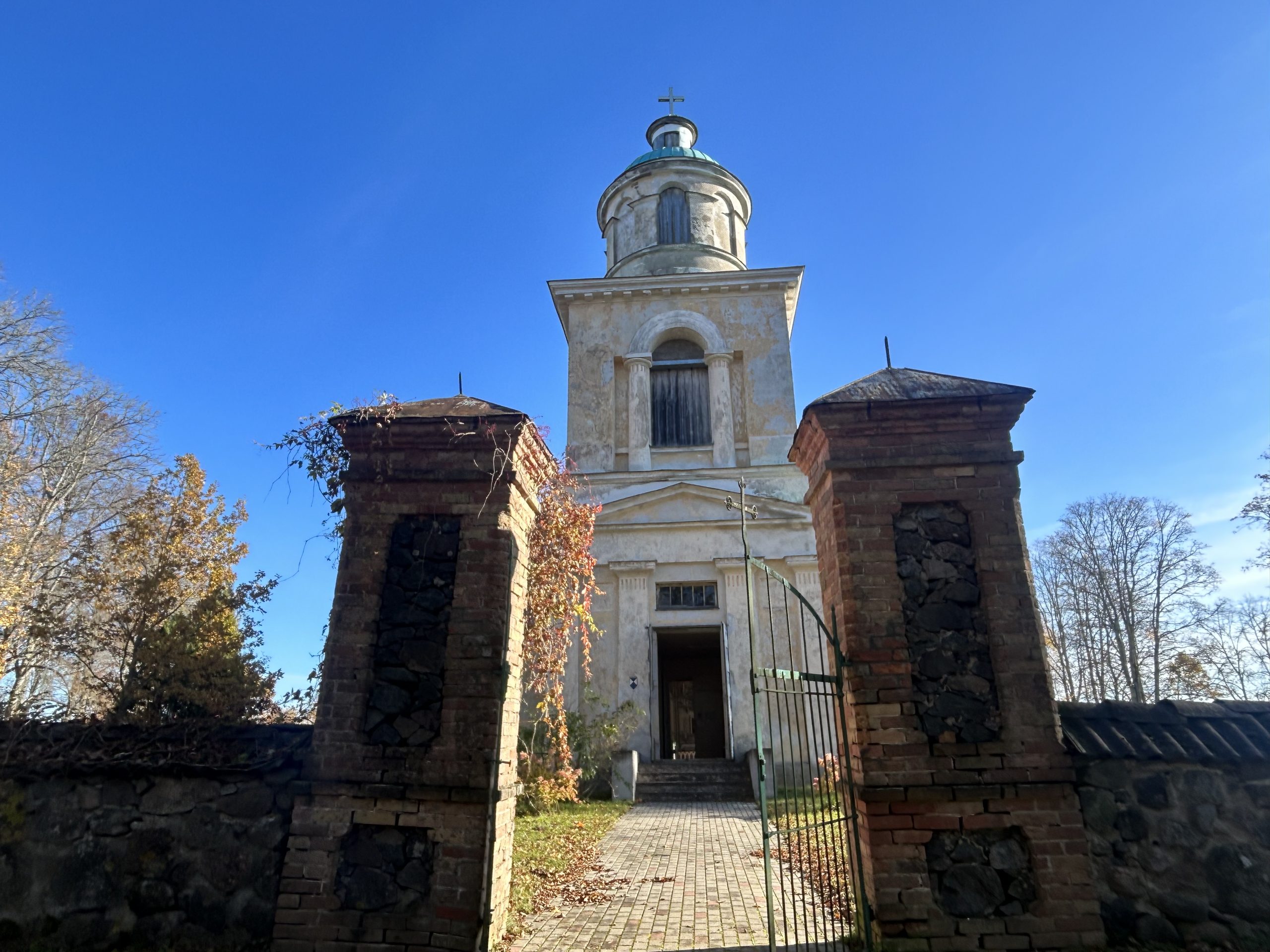 Skats uz Sunākstes (Stendera) baznīcas ieeju caur veciem ķieģeļu vārtiem ar metāla režģi, kas atrodas zem augsta torņa.