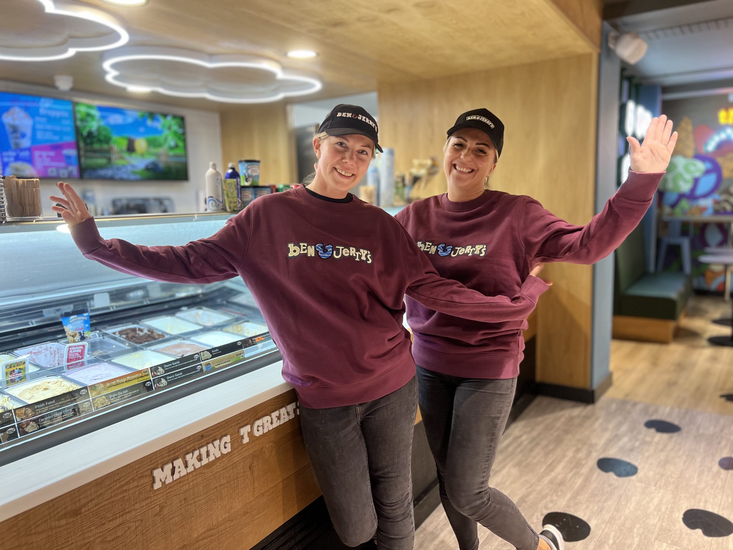 Divas sievietes smaidīgi pozē pie saldējuma letes “Ben & Jerry’s” veikalā.