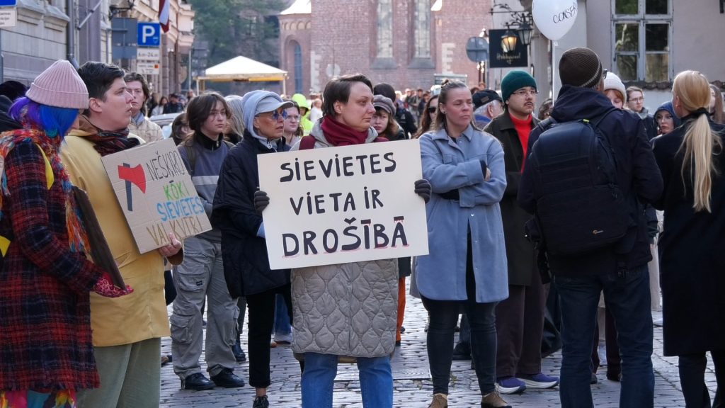 Protesta dalībniece ar plakātu "SIEVETES VIETA IR DROŠĪBĀ".