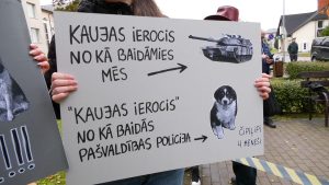 Cilvēks tur plakātu ar uzrakstu “Kaujas ierocis, no kā baidāmies mēs – tanks. ‘Kaujas ierocis’, no kā baidās pašvaldības policija – čipilipi, 4 mēneši”, kur redzams tanks un kucēna attēls.