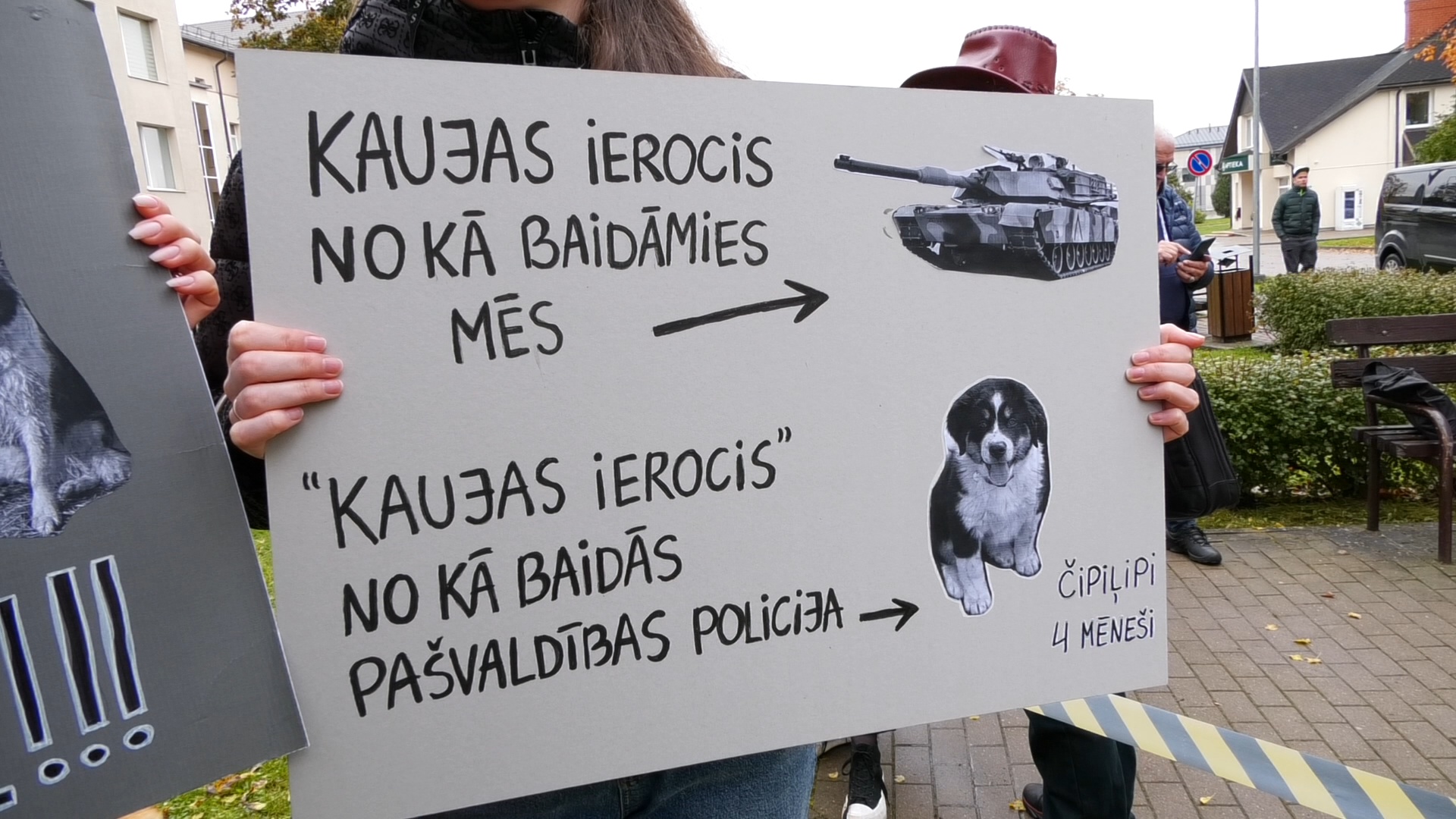Cilvēks tur plakātu ar uzrakstu “Kaujas ierocis, no kā baidāmies mēs – tanks. ‘Kaujas ierocis’, no kā baidās pašvaldības policija – čipilipi, 4 mēneši”, kur redzams tanks un kucēna attēls.