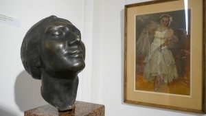 Mākslas izstādē redzama sievietes galvas skulptūra un glezna ar dejotājas portretu.