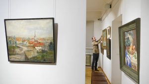 Vīrietis mākslas galerijā pie sienas izkārto gleznas, gatavojot izstādi.