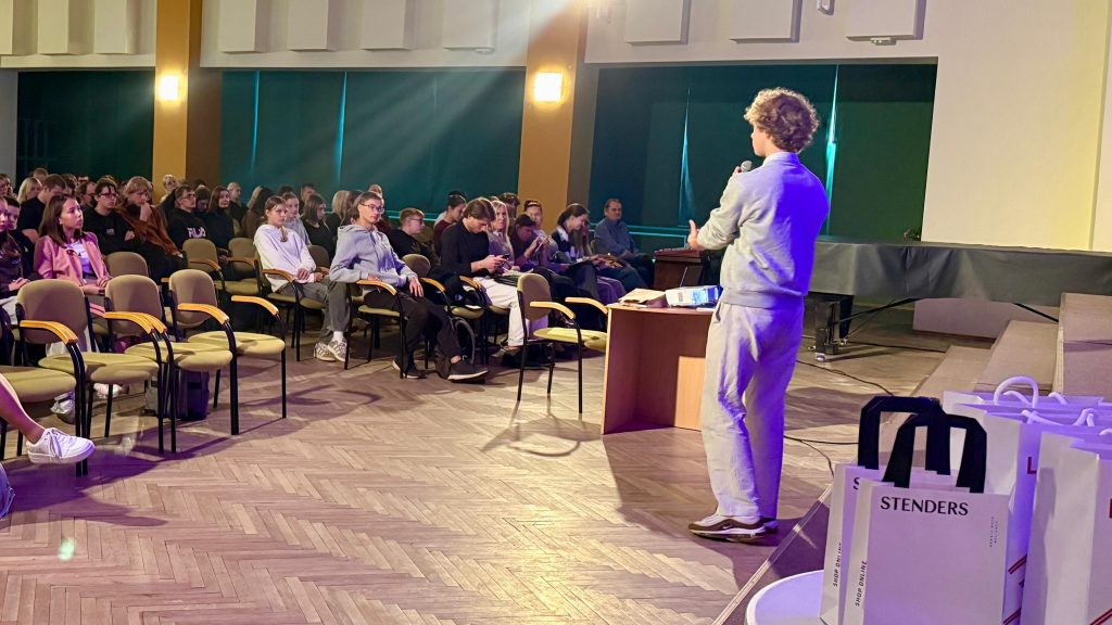 Jauns vīrietis, stāvot uz skatuves, uzrunā sēdošo skolēnu auditoriju.