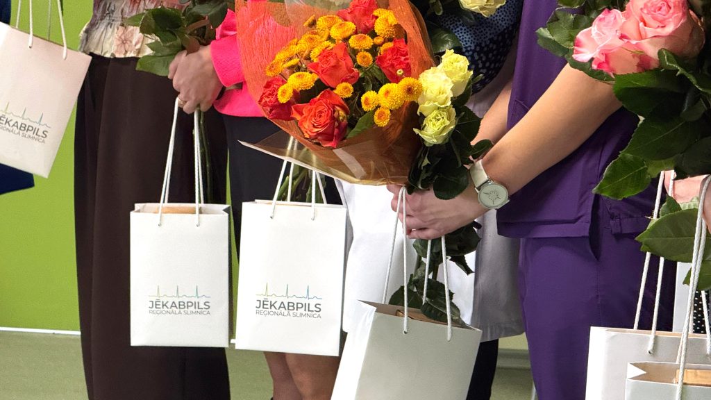 Mediķu rokas, kas tur ziedu pušķus (oranžas rozes, dzeltenas krizantēmas, rozā rozes) un baltus dāvanu maisiņus ar Jēkabpils reģionālās slimnīcas logotipu.