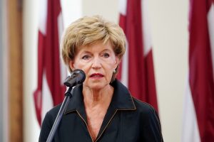 Inese Vaidere runā pie mikrofona Latvijas karogu fonā.