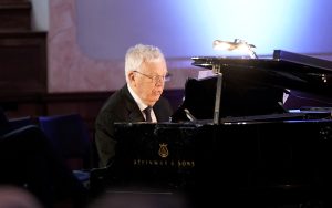Raimonds Pauls melnā uzvalkā spēlē klavieres koncertzālē.