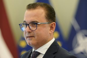 Veselības ministrs Hosams Abu Meri tuvplānā ar brillēm un uzvalkā, fonā Latvijas un ES karogu elementi.