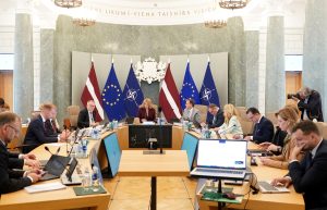 Latvijas Ministru kabineta sēde ar premjerministri centrā, ministriem sēžot pie liela, apaļa galda ar karogiem fonā.