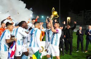 Futbola komanda svin uzvaru stadionā, spēlētāji paceļ čempionu kausu un priecājas.