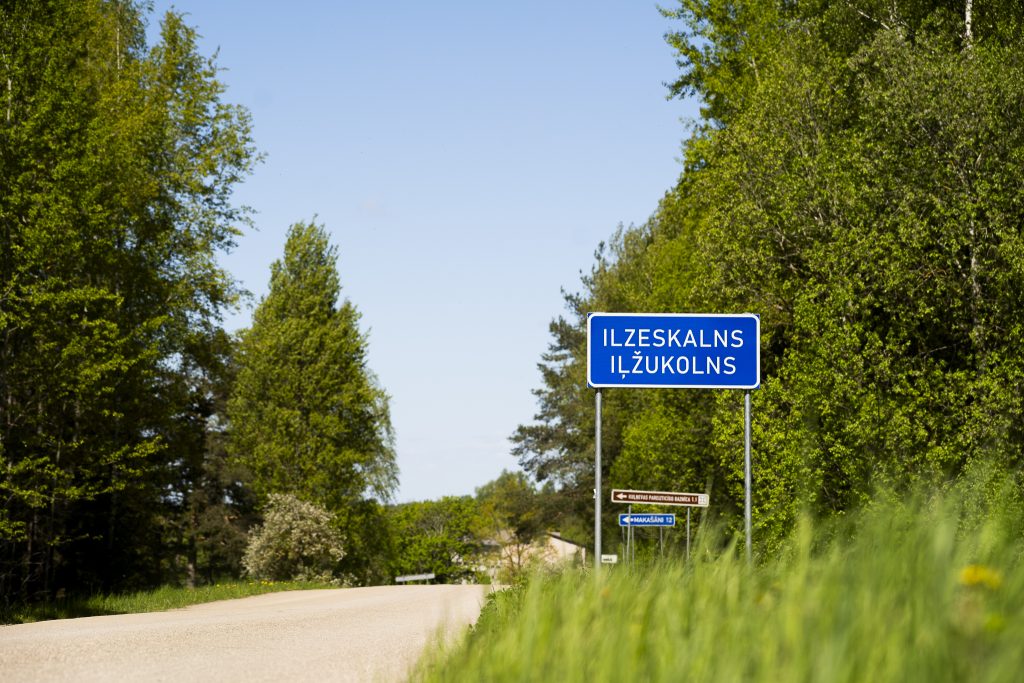 Zila ceļa zīme ar uzrakstu "ILZESKALNS ILŽUKOLNS" uz brūna ceļa pie zaļiem kokiem Nautrēnu kultūrtelpas teritorijā.