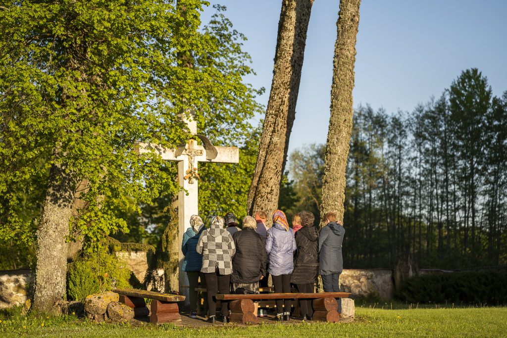 Cilvēki pulcējas pie liela, balta krusta zem kokiem lauku ainavā.