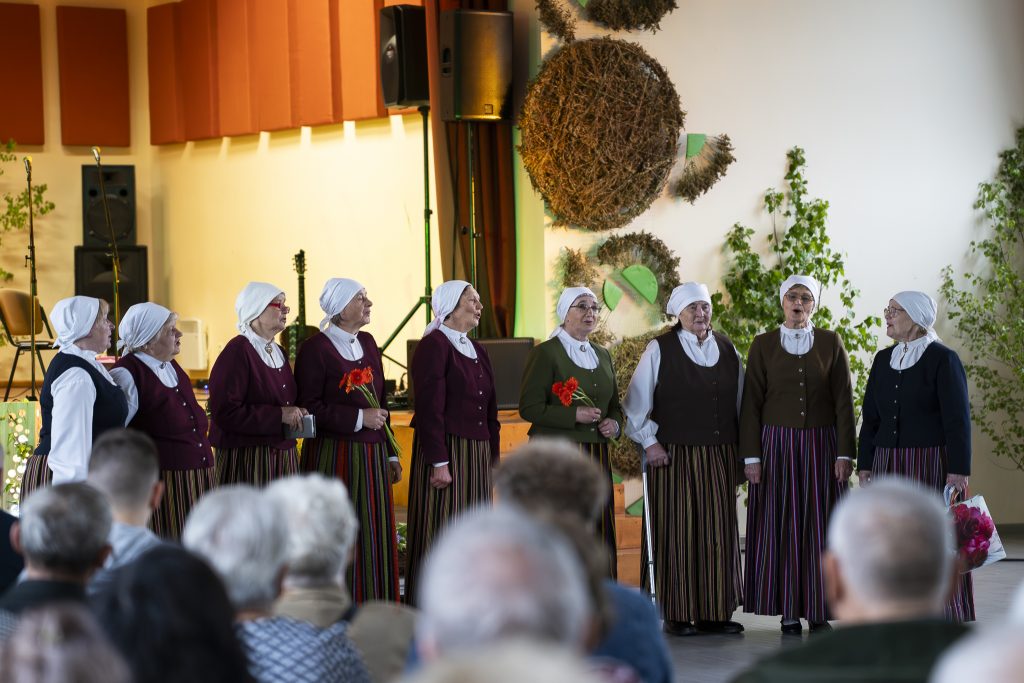 Sievietes tautas tērpos ar baltiem lakatiem dzied folkloras kopa priekšnesumā uz skatuves.