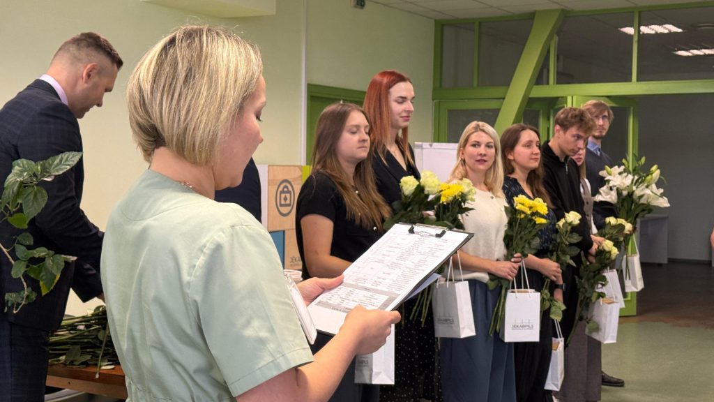 Mediķe gaiši zaļā tērpā nolasot sarakstu, stāv priekšplānā, aiz viņas rindā jauni rezidenti un absolventi ar ziediem un dāvanu maisiņiem.