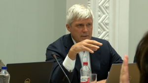 Vīrietis uzstājas domes sēdē, sēžot pie galda ar klēpjdatoru un mikrofonu.