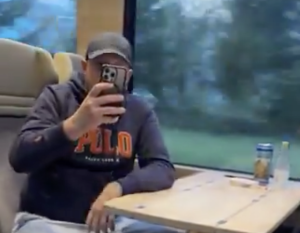 Vīrietis ar cepuri un džemperi sēž vilcienā un fotografē ar telefonu.