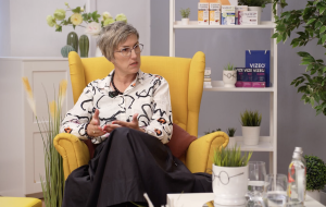 Ilze Maksima sēž dzeltenā krēslā un runā studijas vidē ar augiem un produktiem fonā.