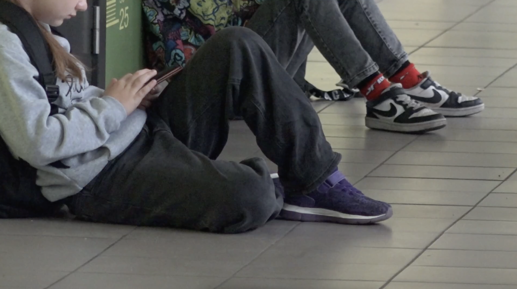 Tuvplāns skolēnam, kurš sēž uz skolas grīdas un lieto mobilo tālruni, fonā redzamas vēl divas kājas.