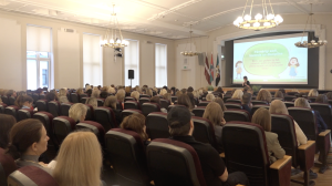 Plaša konferenču zāle ar simtiem klausītāju, kas sēž krēslos un skatās uz skatuvi, kur notiek prezentācija par bērna disciplīnu.