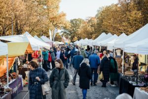 Plašs skats uz Simjūda gadatirgu Valmierā, kur cilvēki pulcējas starp baltiem tirdzniecības telšu jumtiem rudenīgā ielā.