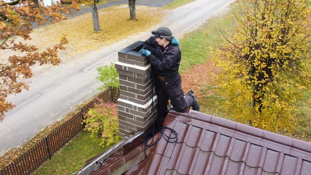 Skursteņslauķe Līga Ondzule tīra mūra skursteni uz mājas jumta rudenīgā ainavā, ar dzeltenām lapām visapkārt.