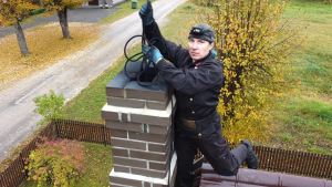 Skursteņslauķe Līga Ondzule tradicionālā, tumšā darba apģērbā, ar pieres lukturīti, sēžot uz sarkanbrūna jumta pie ķieģeļu skursteņa un tur virvi.