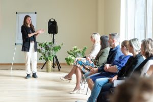 Sieviete vada darbnīcu, stāvot pie tāfeles un runājot ar auditoriju Tukumā.