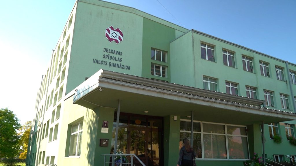 Jelgavas Spīdolas Valsts ģimnāzijas ēkas fasāde maigi zaļā krāsā ar uzrakstu un skolas ģerboni saulainā dienā.