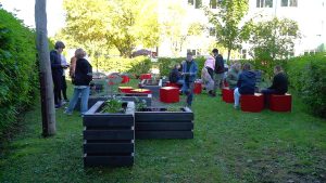Plašs skats uz Jelgavas Spīdolas Valsts ģimnāzijas āra klasi ar skolēniem, kas sēž uz sarkaniem kubiem un strādā pie melnām dobēm.