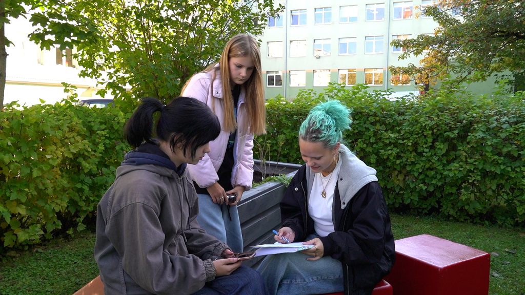 Trīs skolnieces, viena ar tirkīza matiem, sēž un strādā ar pierakstiem un telefonu Jelgavas Spīdolas Valsts ģimnāzijas āra mācību klasē.