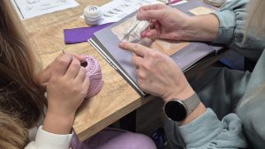 Sieviete un meitene rokdarbu nodarbībā apgūst tamborēšanas tehniku ar smalku dziju un āķi.