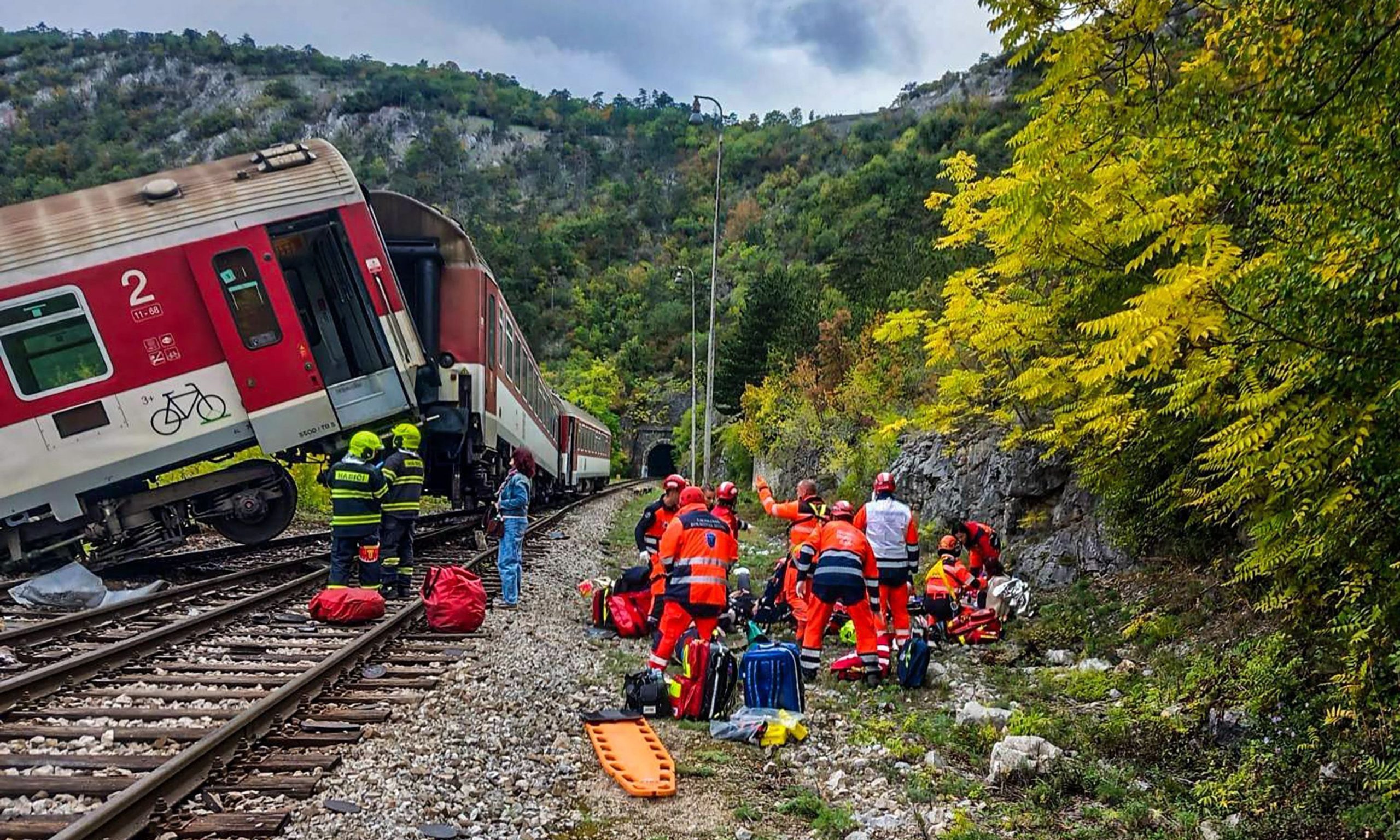 Vilciena vagons, kas nobraucis no sliedēm kalnainā apvidū, un glābēji oranžos tērpos pie tuneļa ieejas.