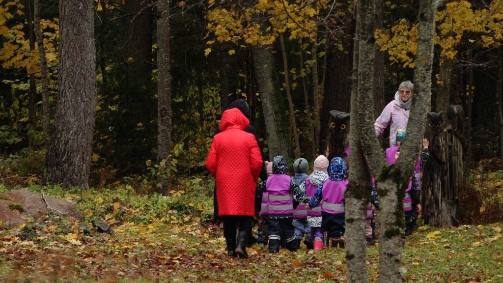 Bērni un divas pieaugušās dodas pastaigā pa rudens lapām noklātu meža taku. Bērni ģērbušies košās vestēs.