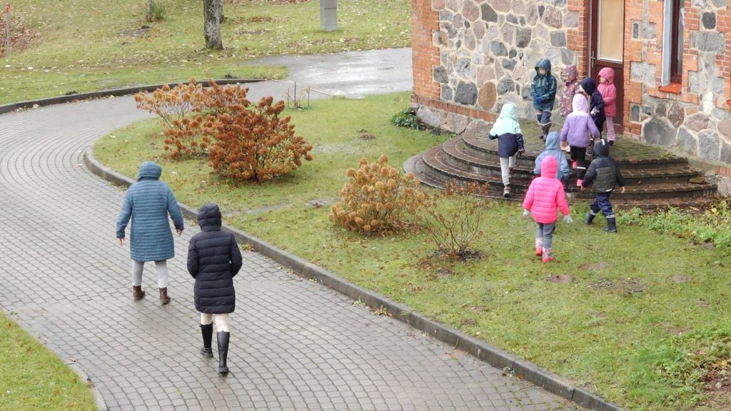 Bērni un divas pieaugušās iet pa bruģētu celiņu un kāpj pa akmens pakāpieniem uz Sējas pamatskolas ēkas pusi. Visi tērpušies mēteļos un lietus jakās.