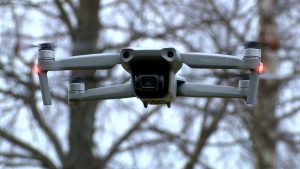 Pelēks drons “Mavic Air 2” lido gaisā ar ieslēgtām gaismām, fonā koki.