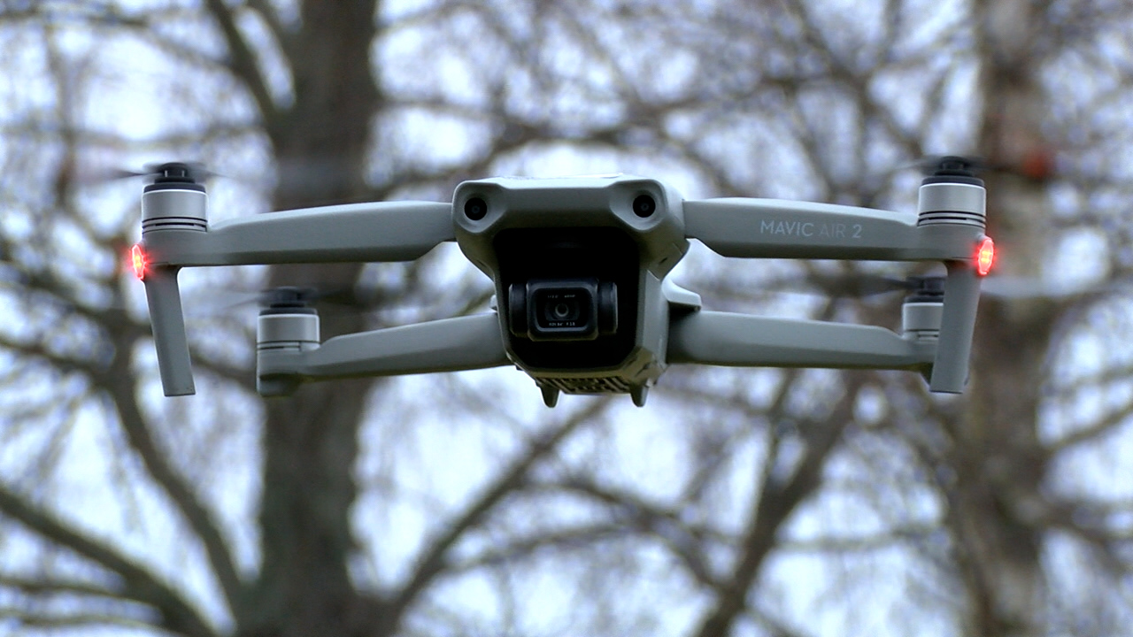 Pelēks drons “Mavic Air 2” lido gaisā ar ieslēgtām gaismām, fonā koki.