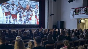 Pilna kinozāle ar skatītājiem, kas vēro uz lielā ekrāna redzamo hokeja komandas “Sēļi” spēlētāju koptēlu dokumentālās filmas "No dīķa līdz uzvarām" pirmizrādes laikā.