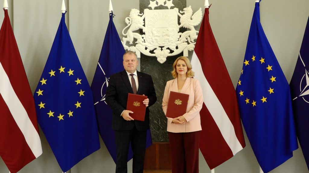 Ministru prezidente Evika Siliņa un LPS priekšsēdētājs Gints Kaminskis stāv starp Latvijas, ES un NATO karogiem ar sarkanām mapēm rokās.